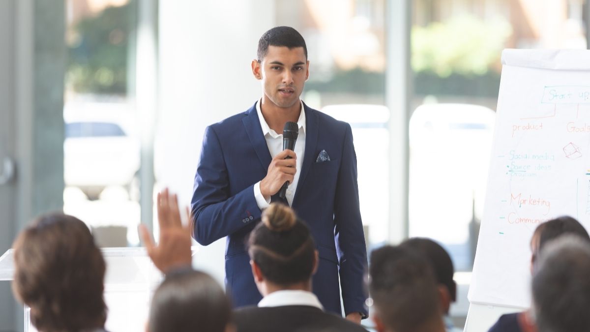 Charismatic man in a suit holding a microphone, standing in front of a seated audience as he prepares to speak